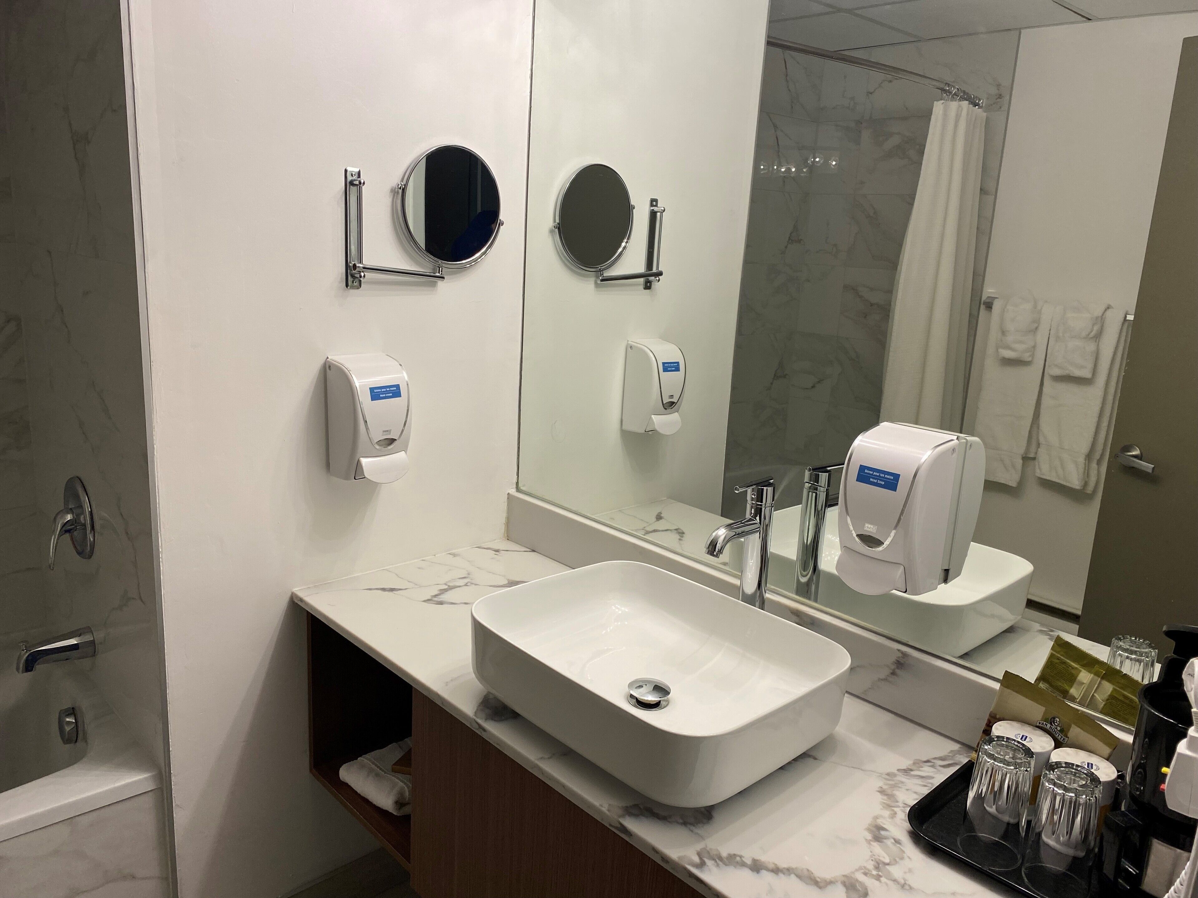 A bathroom with a large mirror, two round mirrors, a white sink, a shower area, and a towel rack with towels.