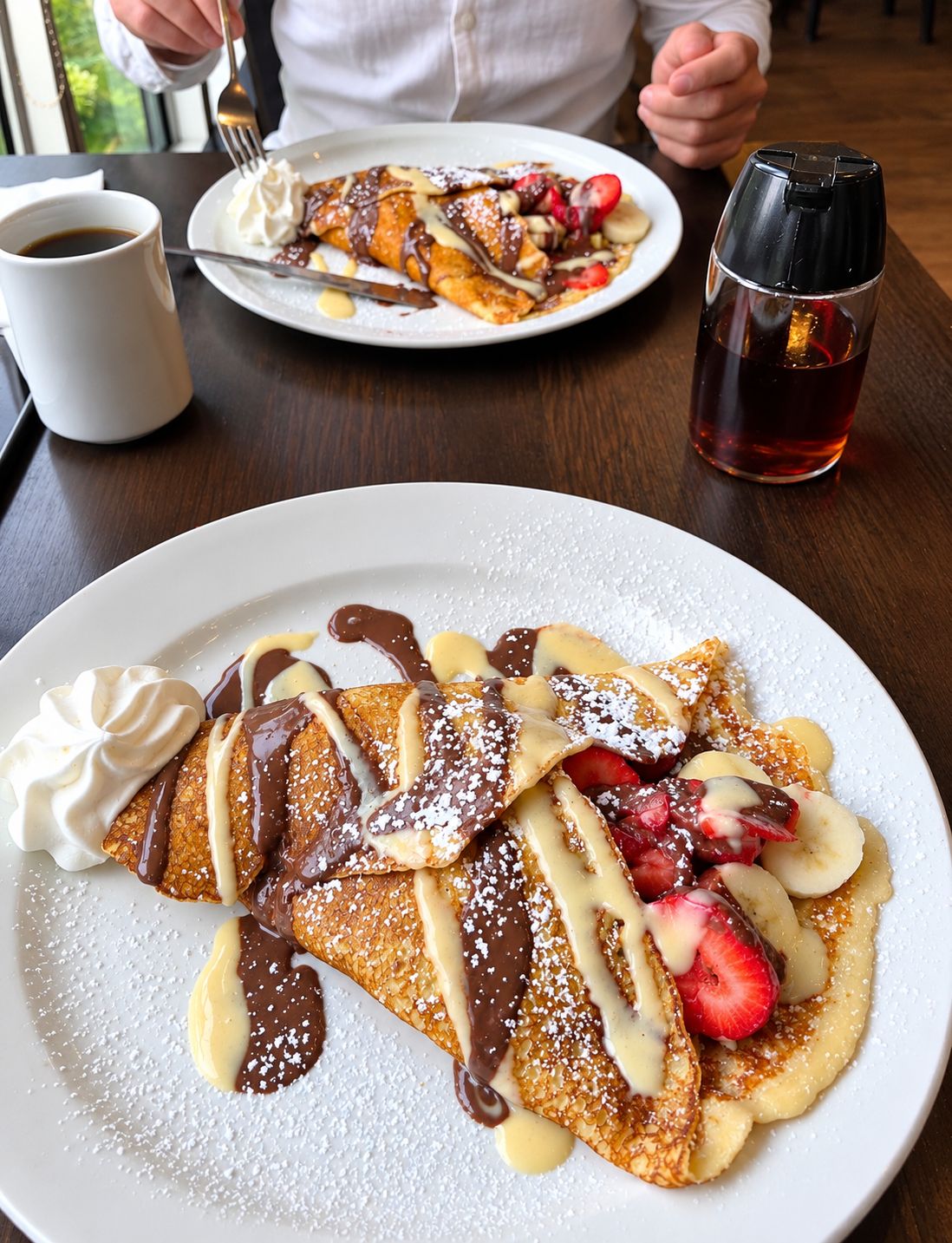 Un plato de panqueques con crema batida, salsa de chocolate y fruta, sobre una mesa con una taza de café y un vaso de té.