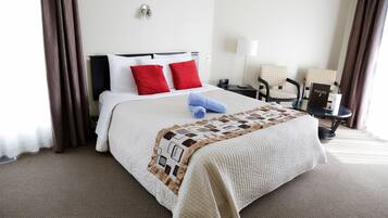 Una cama cuidadosamente tendida con almohadas rojas y blancas, una toalla azul y un edredón acolchado. Junto a la cama hay una mesita con una lámpara y un libro titulado