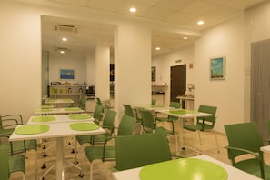 A restaurant with green chairs and white tables, a kitchen area in the background, and a painting on the wall.