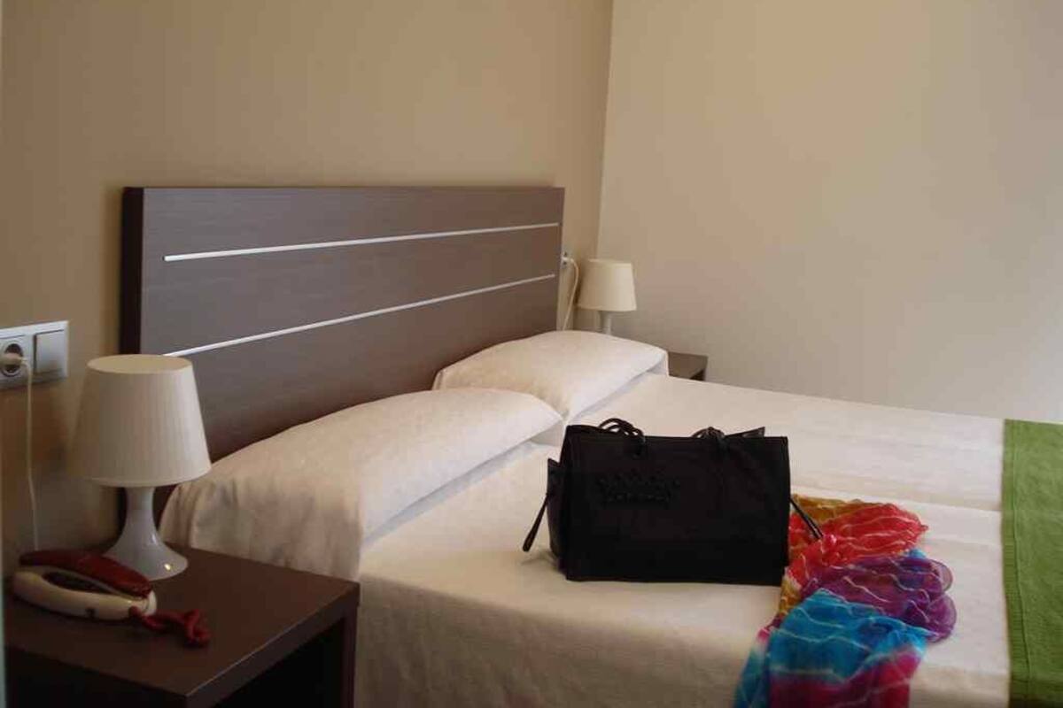 A hotel room with a bed, a nightstand with a lamp, and a black bag on the bed.