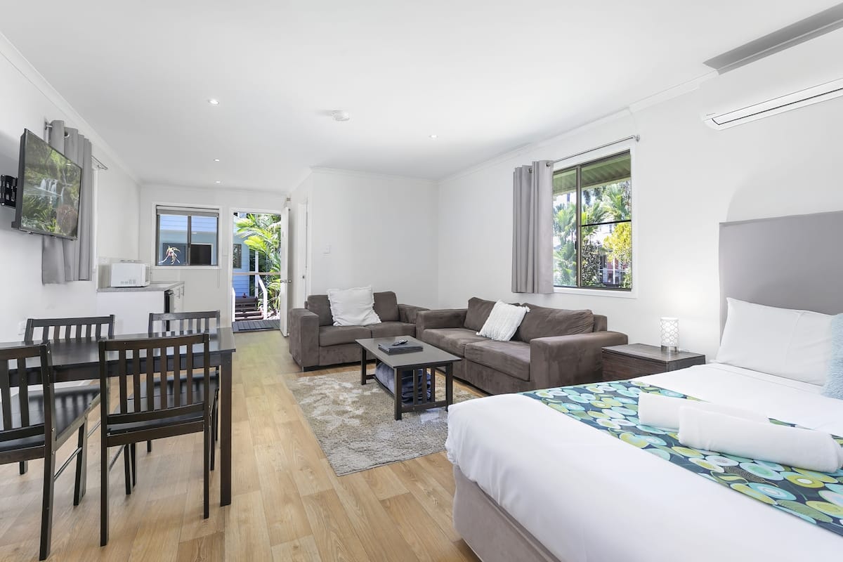 A modern hotel room with a bed, a sofa, a dining table with chairs, and a small kitchen area.