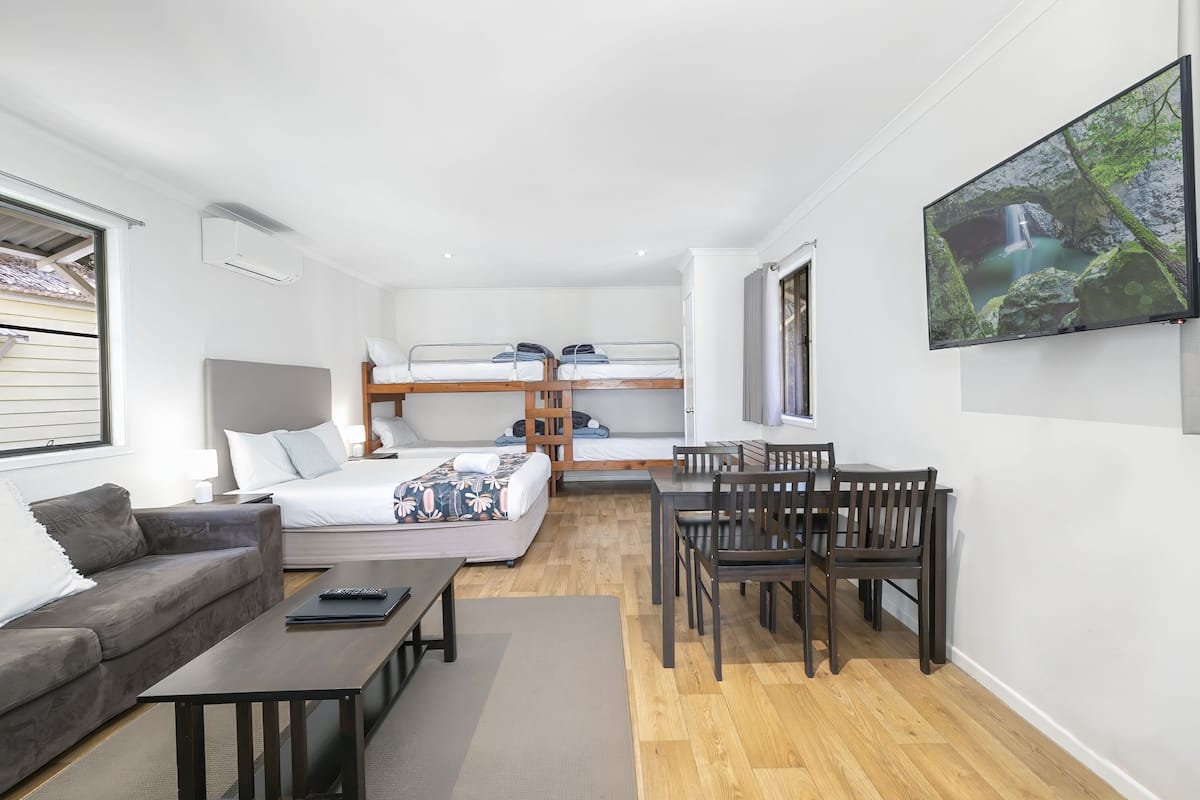 A modern, well-lit room with a sofa, a dining table with chairs, a bunk bed, and a flat-screen TV mounted on the wall.