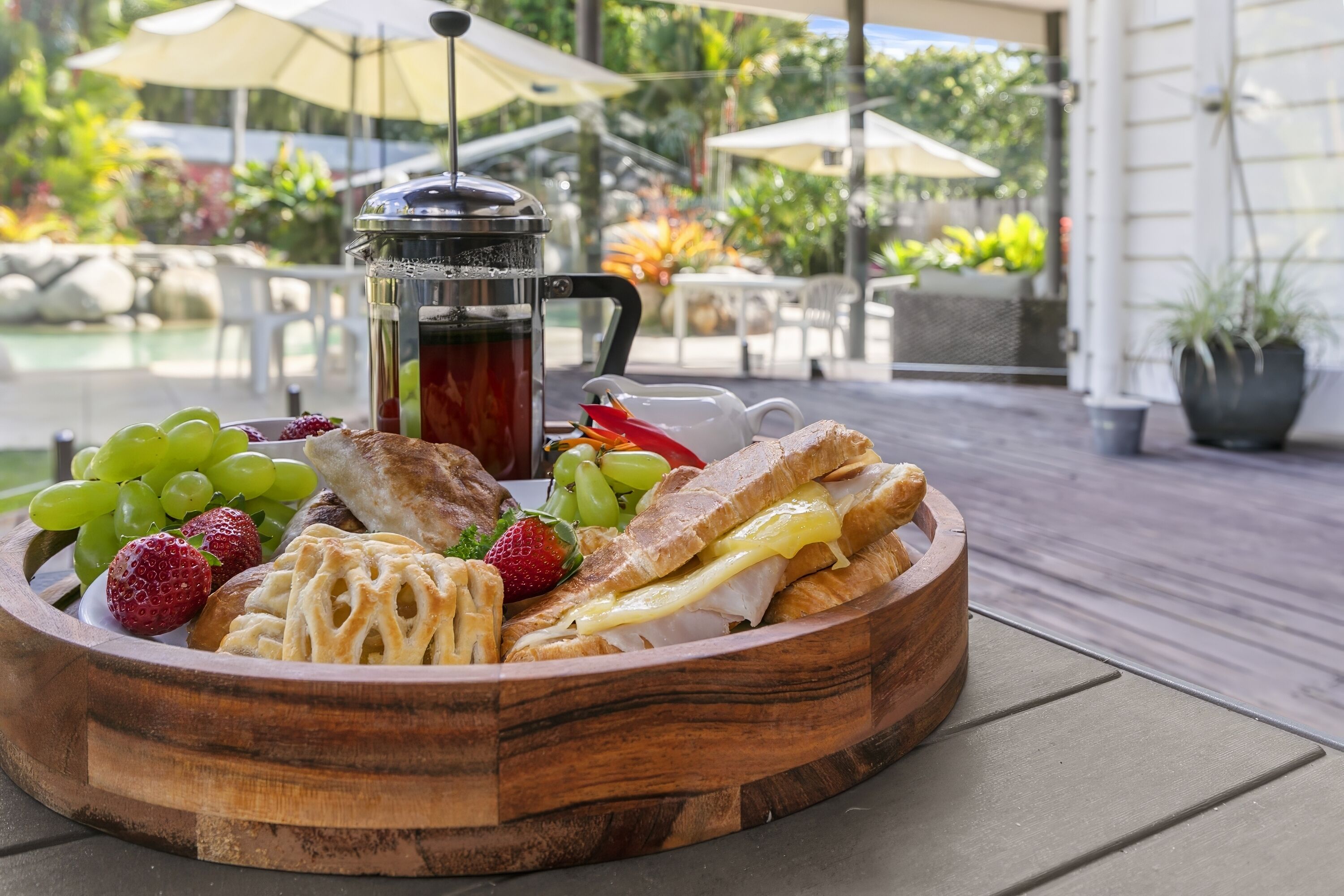 A wooden tray with sandwiches, grapes, strawberries, and a coffee pot on an outdoor patio.