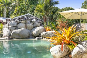 A pool area with a stone wall, tropical plants, and a wooden fence. - Mossman Resort Holiday Villas (Mossman)