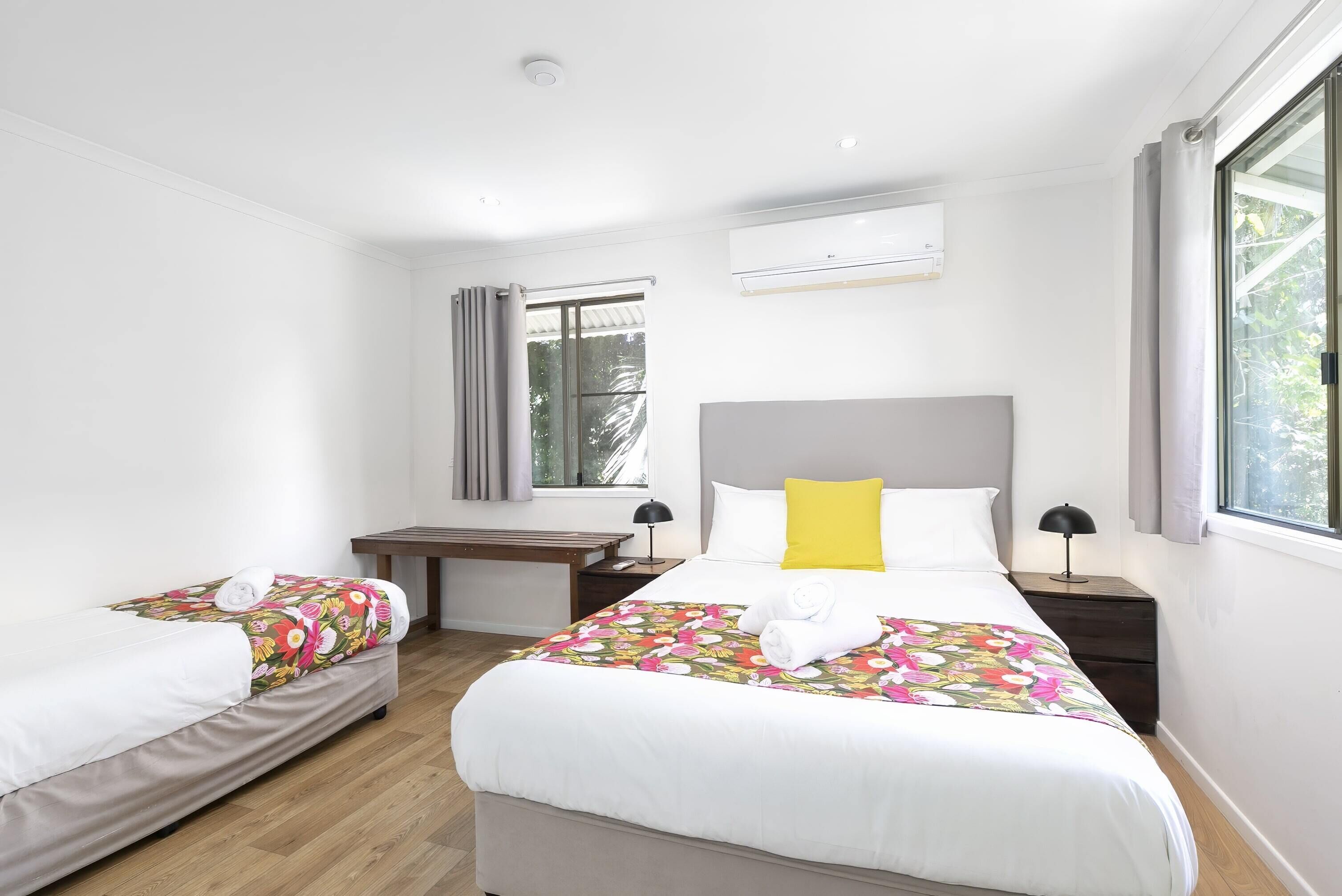 A hotel room with two beds, a desk, a chair, an air conditioning unit, and a window with curtains.