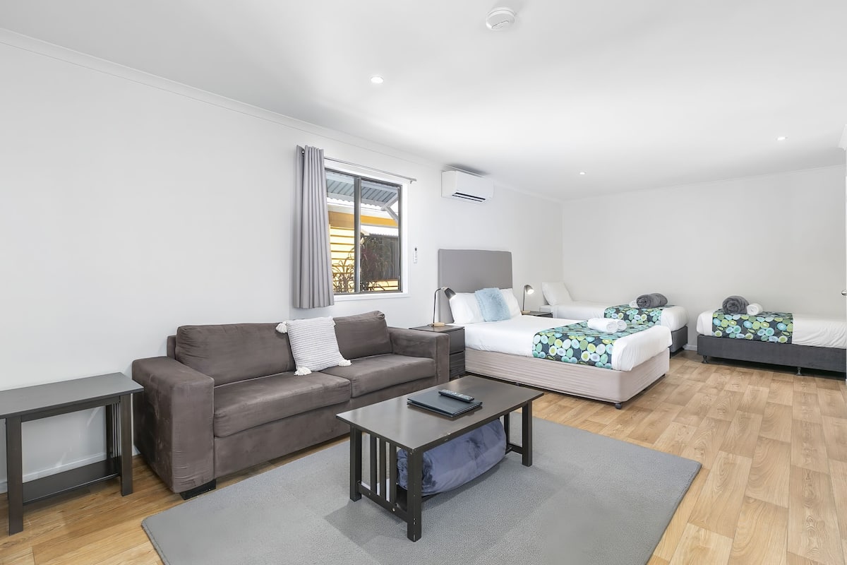 A modern, well-lit room with a sofa, a small table, a bed with a patterned bedspread, and a window with curtains.