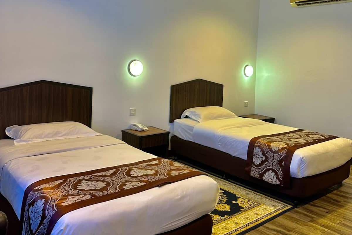 A hotel room with two beds, each with a wooden headboard, a bedside table with a lamp, and a wall-mounted air conditioner.