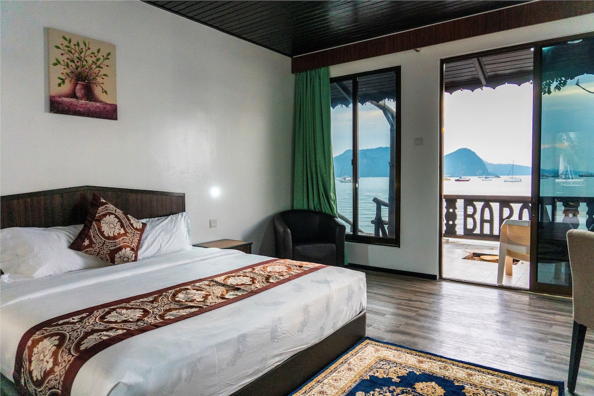 A hotel room with a large bed, a view of the sea, and a balcony with chairs.