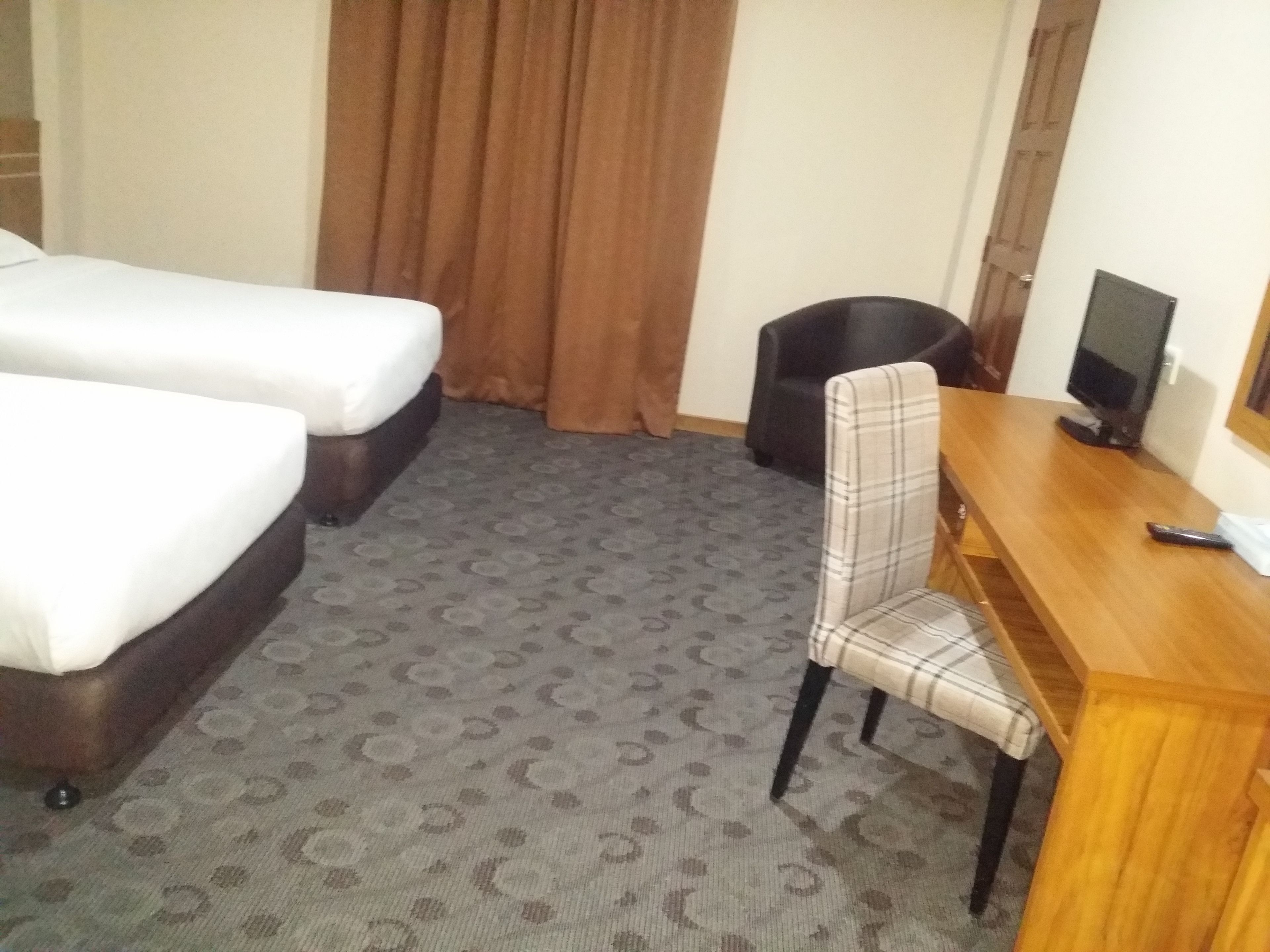 A hotel room with two single beds, a wooden desk, a chair, a television, and a patterned carpet.