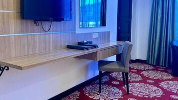 A hotel room with a wooden desk, a flat-screen TV mounted on the wall, a window with curtains, and a red patterned carpet.