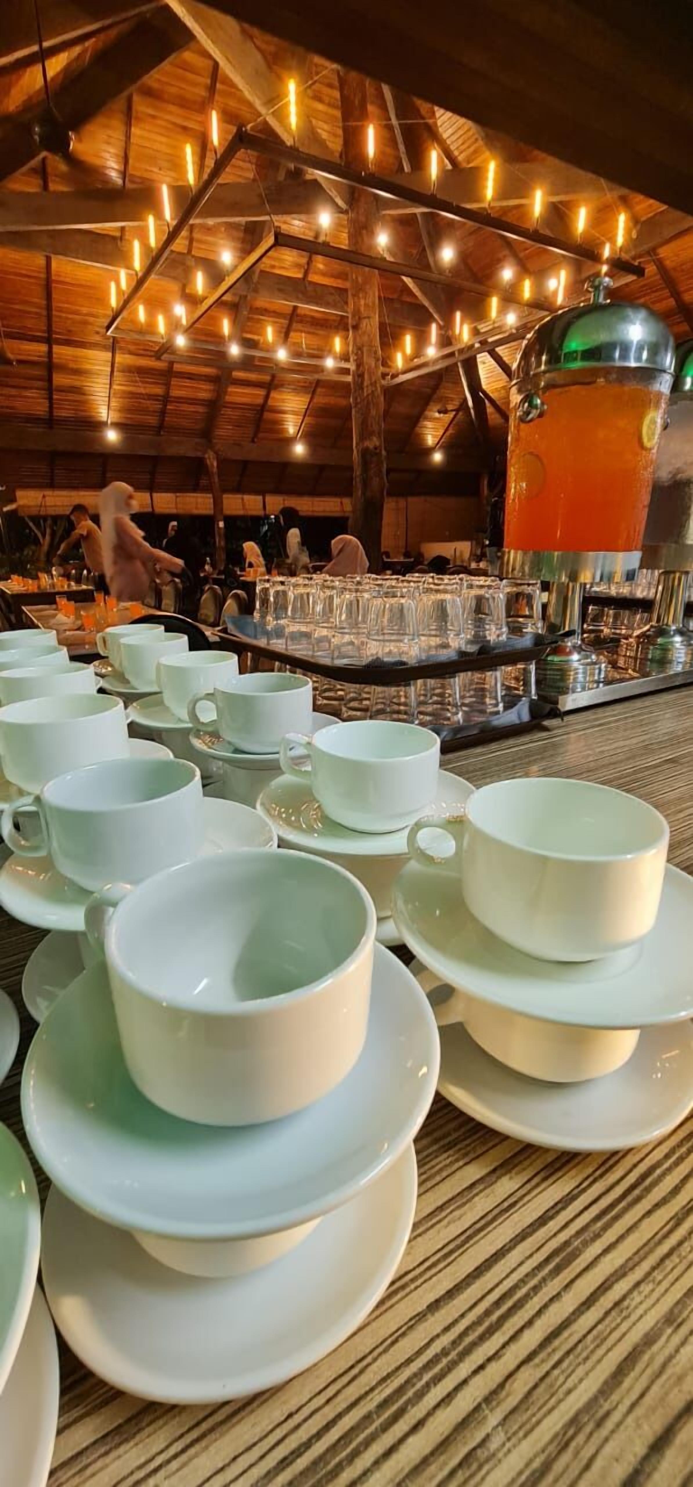 A rustic dining area with wooden beams and string lights, a large beverage dispenser, and neatly arranged white cups and saucers.