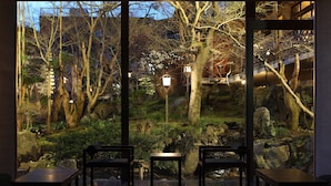 A room with a view of a landscaped garden, featuring a pond, rocks, and trees. There are three tables and chairs arranged for seating. - Ryokan Nenrinbo (Kyoto)