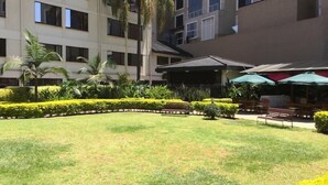 A courtyard with a lawn, palm trees, and a covered seating area with umbrellas. - Jacaranda (Nairobi)