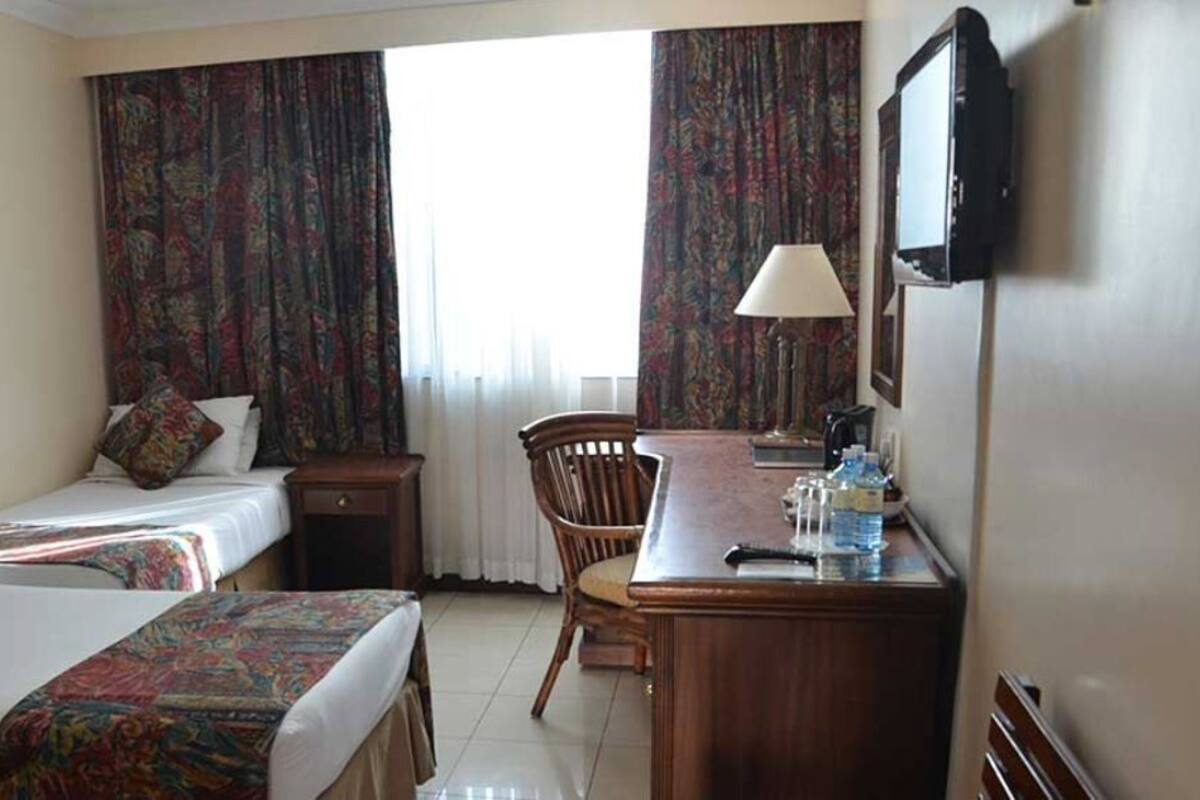 A hotel room with two beds, a desk with a lamp, a chair, and a TV mounted on the wall.