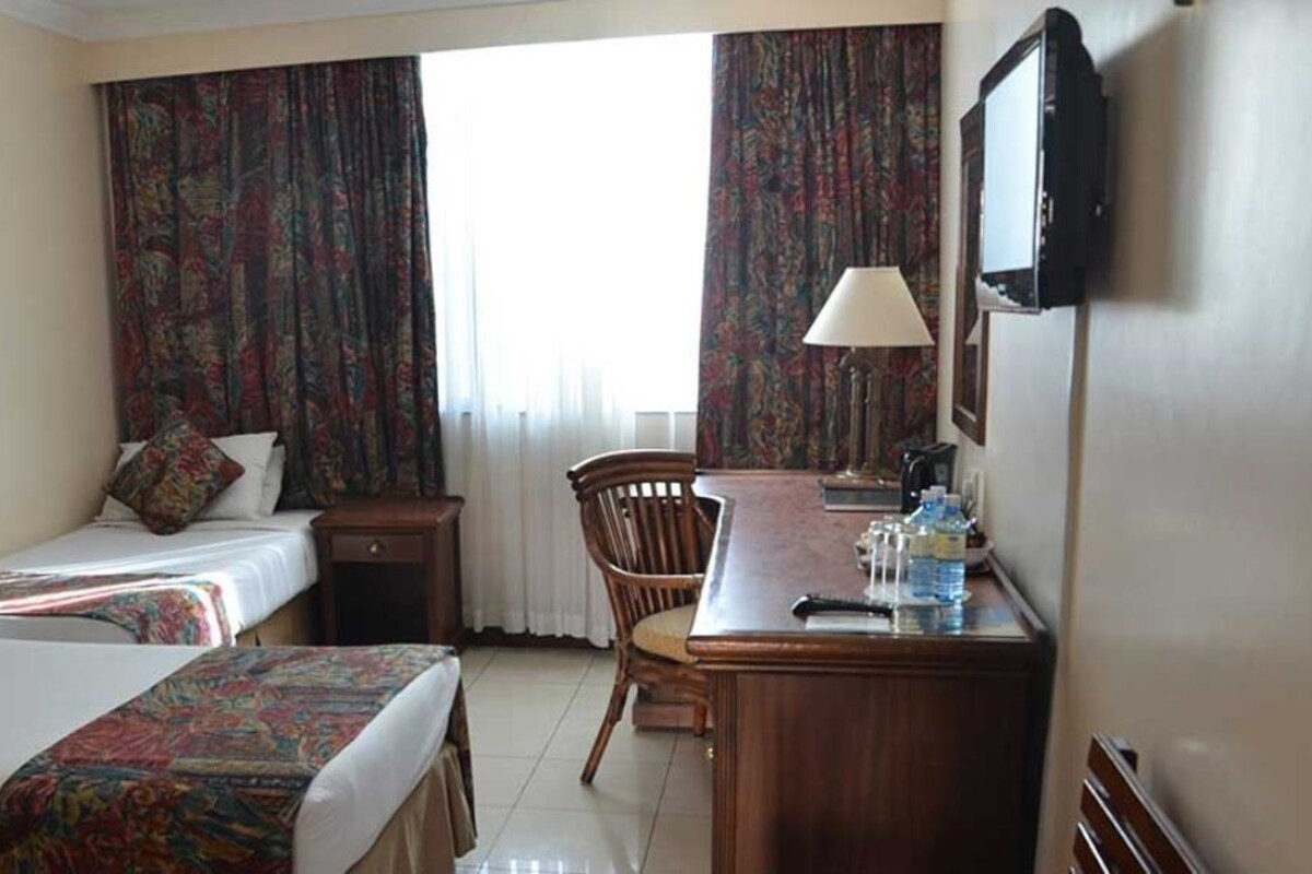 A hotel room with two beds, a desk with a lamp, a chair, and a TV mounted on the wall.