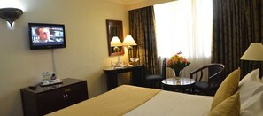 A hotel room with a bed, a desk, a chair, a TV, and a window with curtains. - Jacaranda (Nairobi)