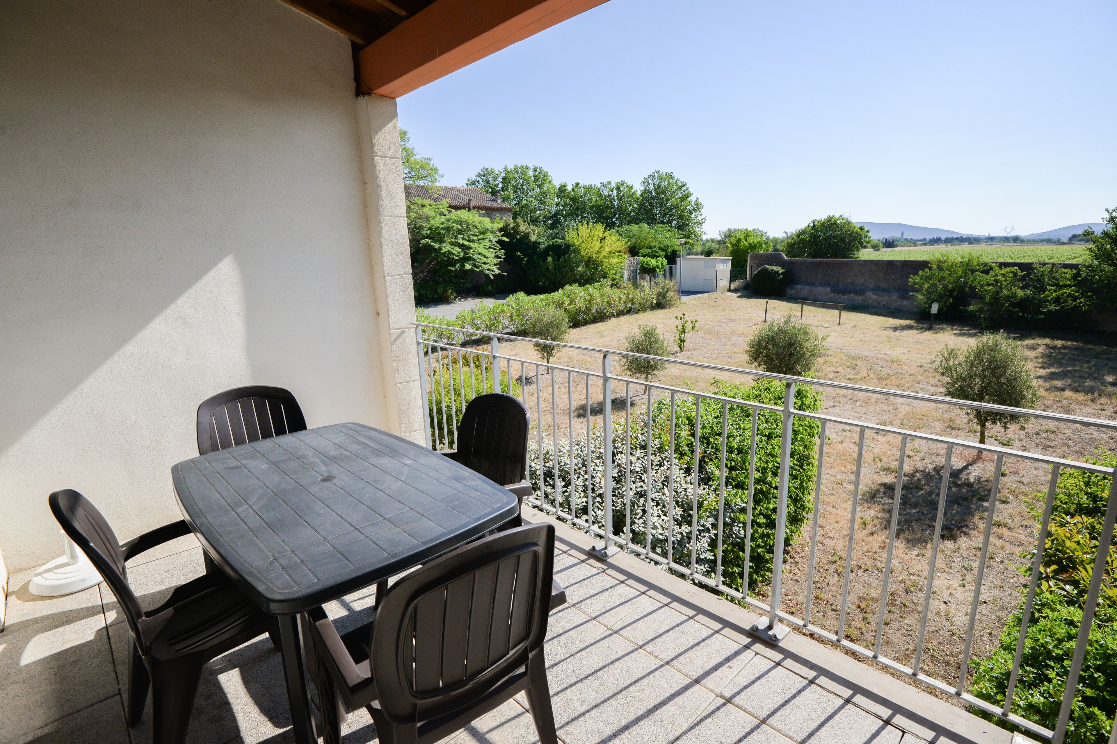 Varanda com mesa e cadeiras, corrimão branco e vista para um jardim e colinas ao longe.