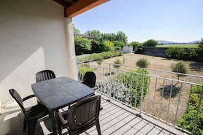 Un balcón con una mesa y sillas, baranda blanca y vistas a un jardín y colinas lejanas.