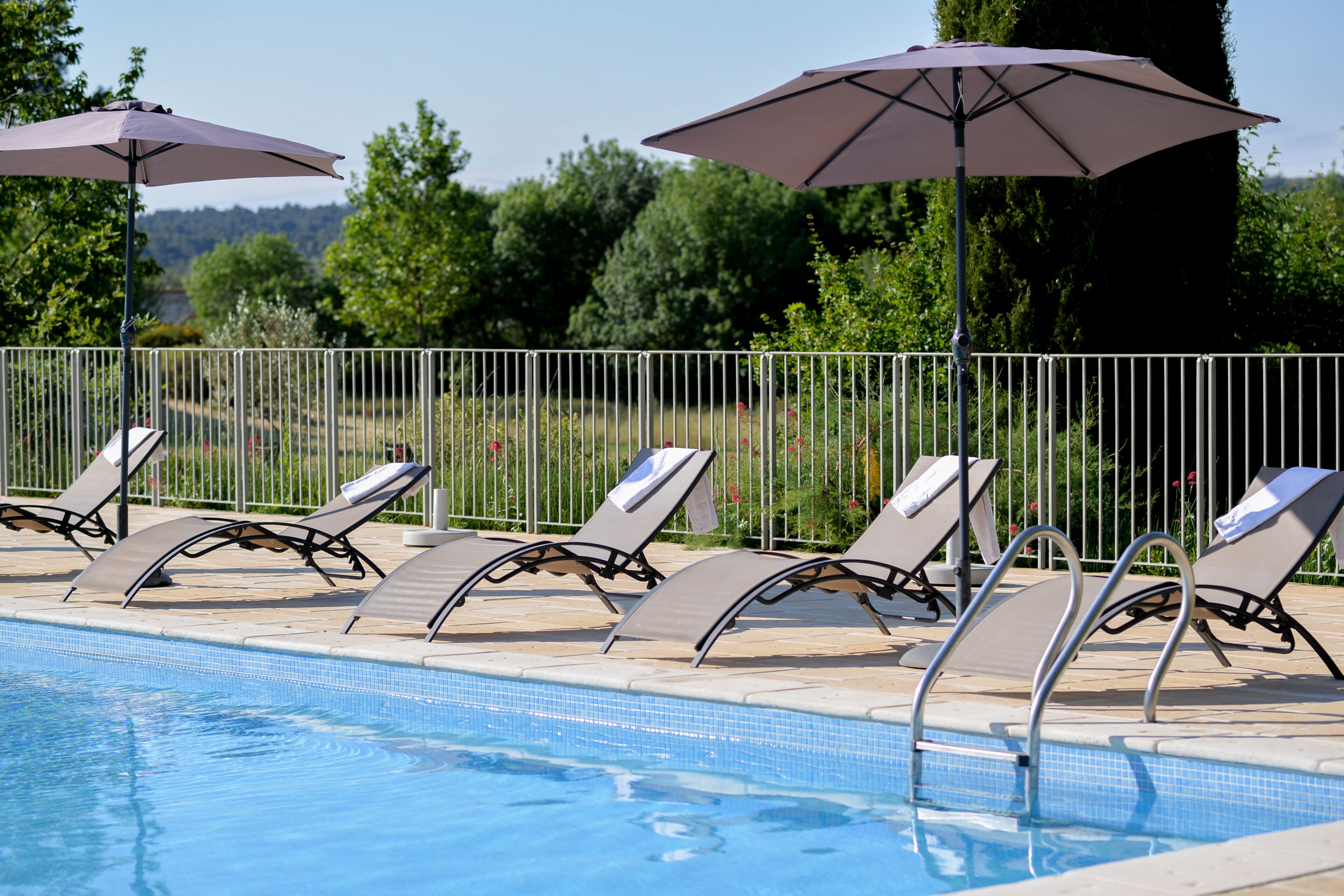A pool area with lounge chairs and umbrellas, surrounded by a metal fence and greenery.