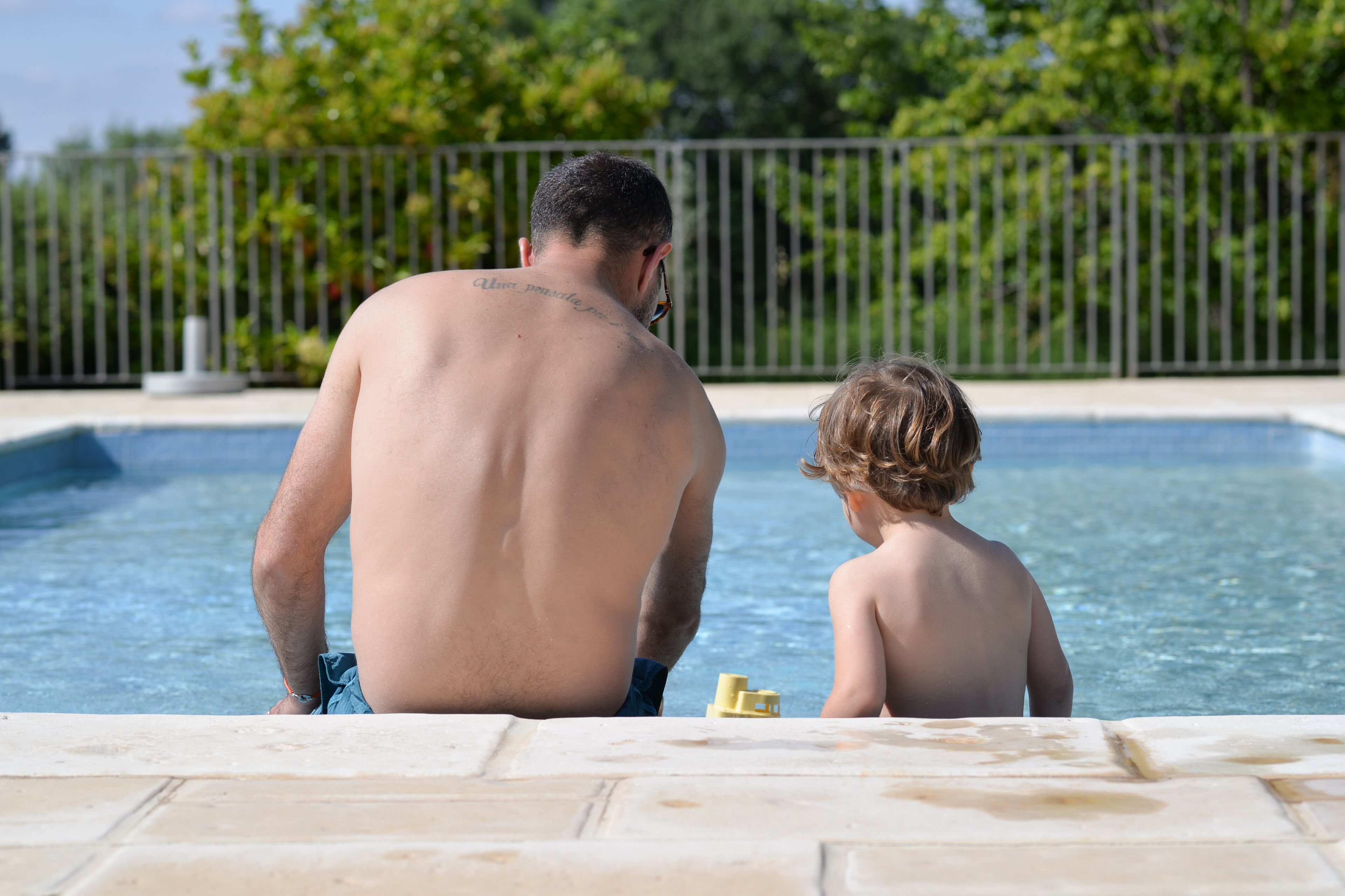 Un homme et un enfant sont assis au bord d’une piscine, avec une clôture et de la végétation en arrière-plan.