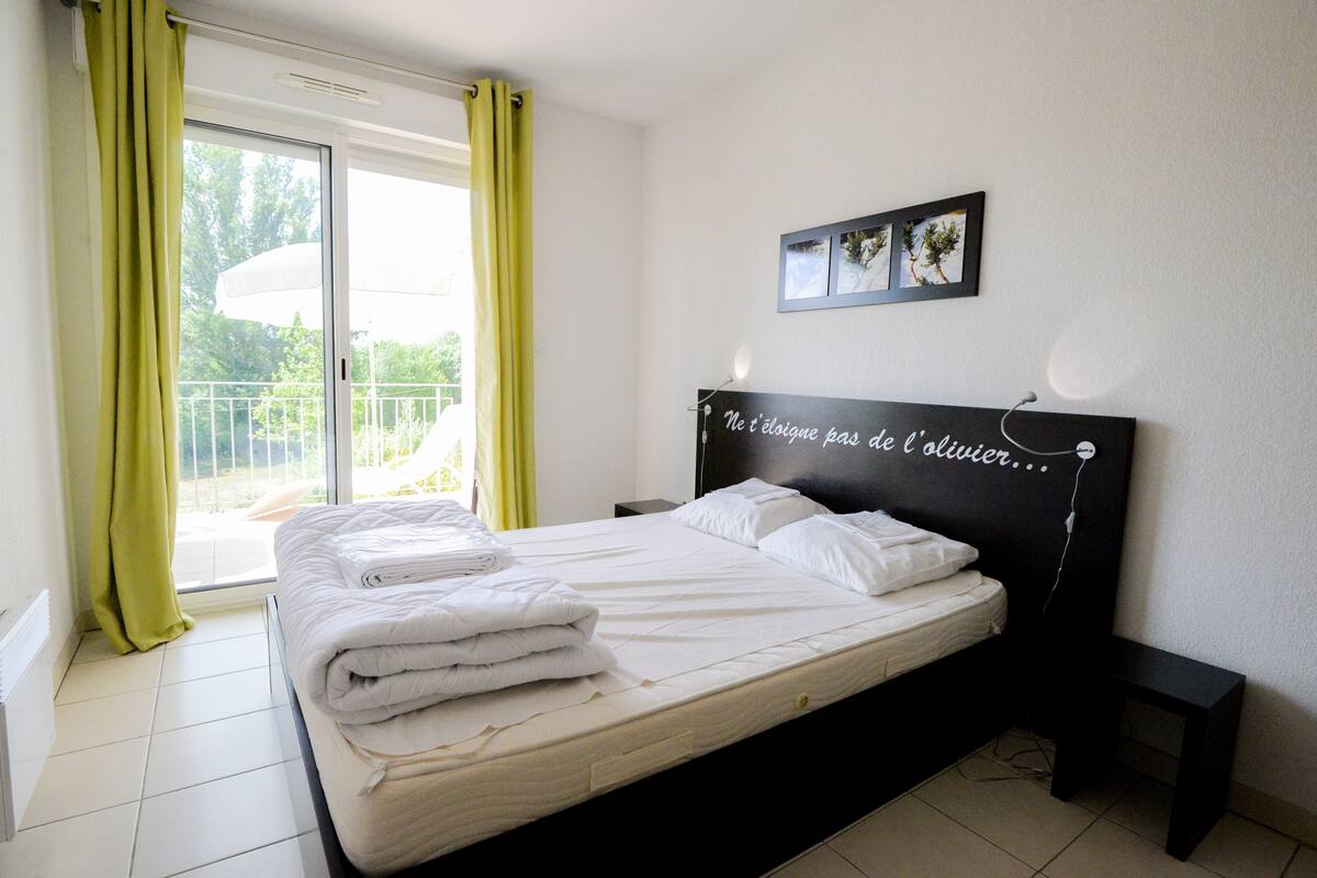 A neatly made bed with white linens and pillows, a black headboard with a quote, a bedside table with a lamp, and a view of greenery through a sliding door.