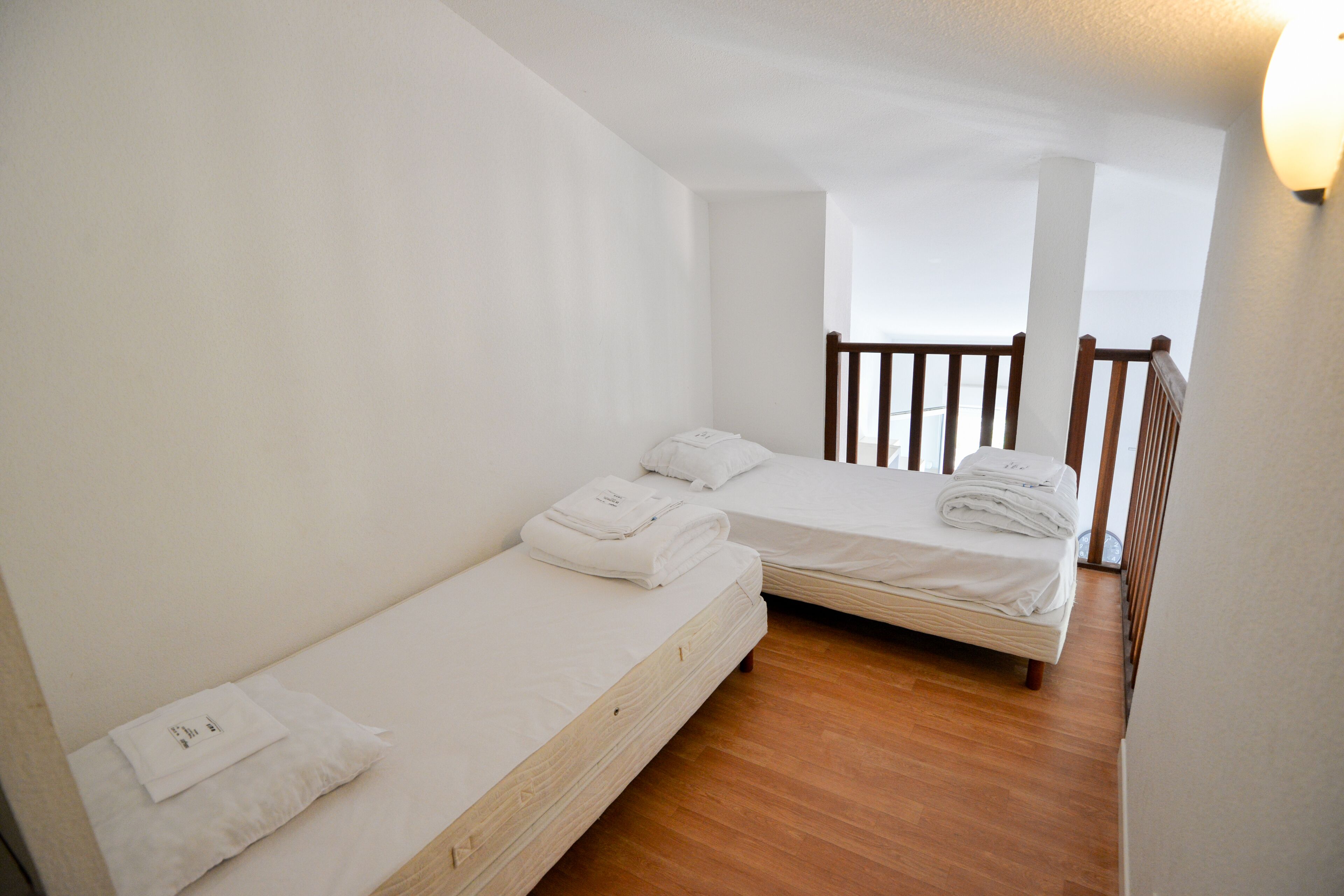 A small, clean room with two single beds, wooden flooring, and a balcony with a railing.