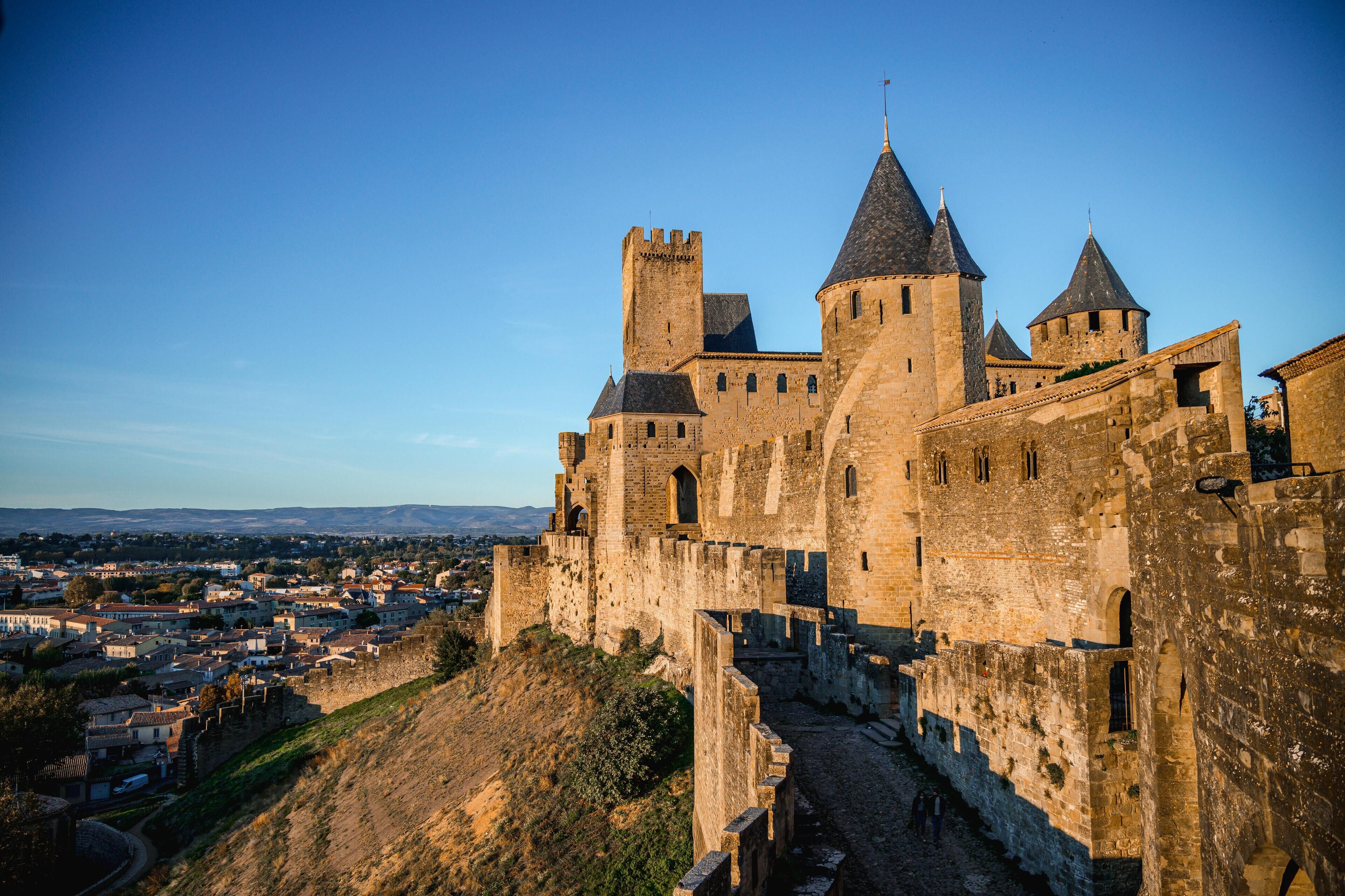 A historic stone castle with multiple towers and a fortified wall, overlooking a town below.