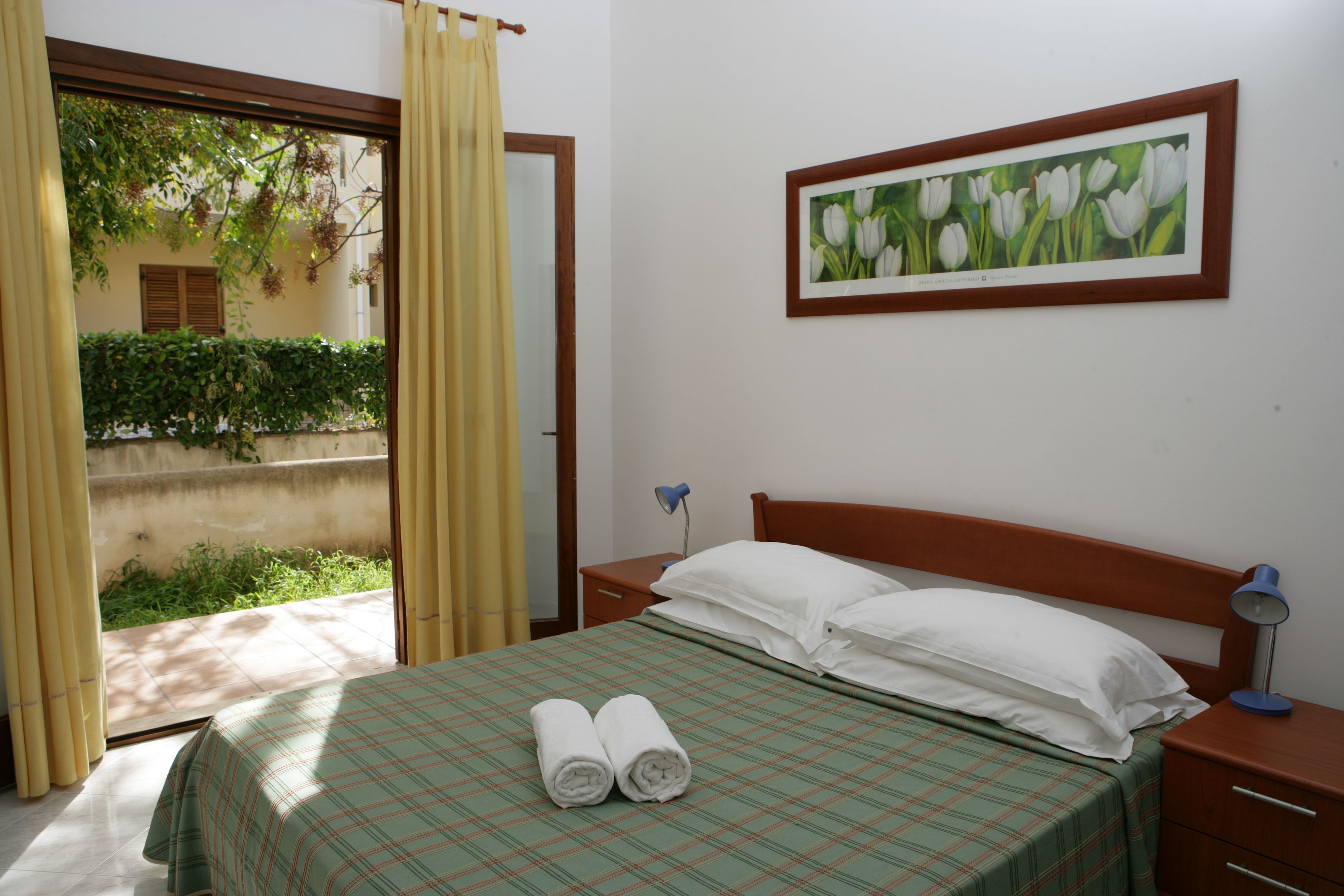 A bedroom with a bed, pillows, a bedside lamp, a framed picture, and a view of greenery outside.