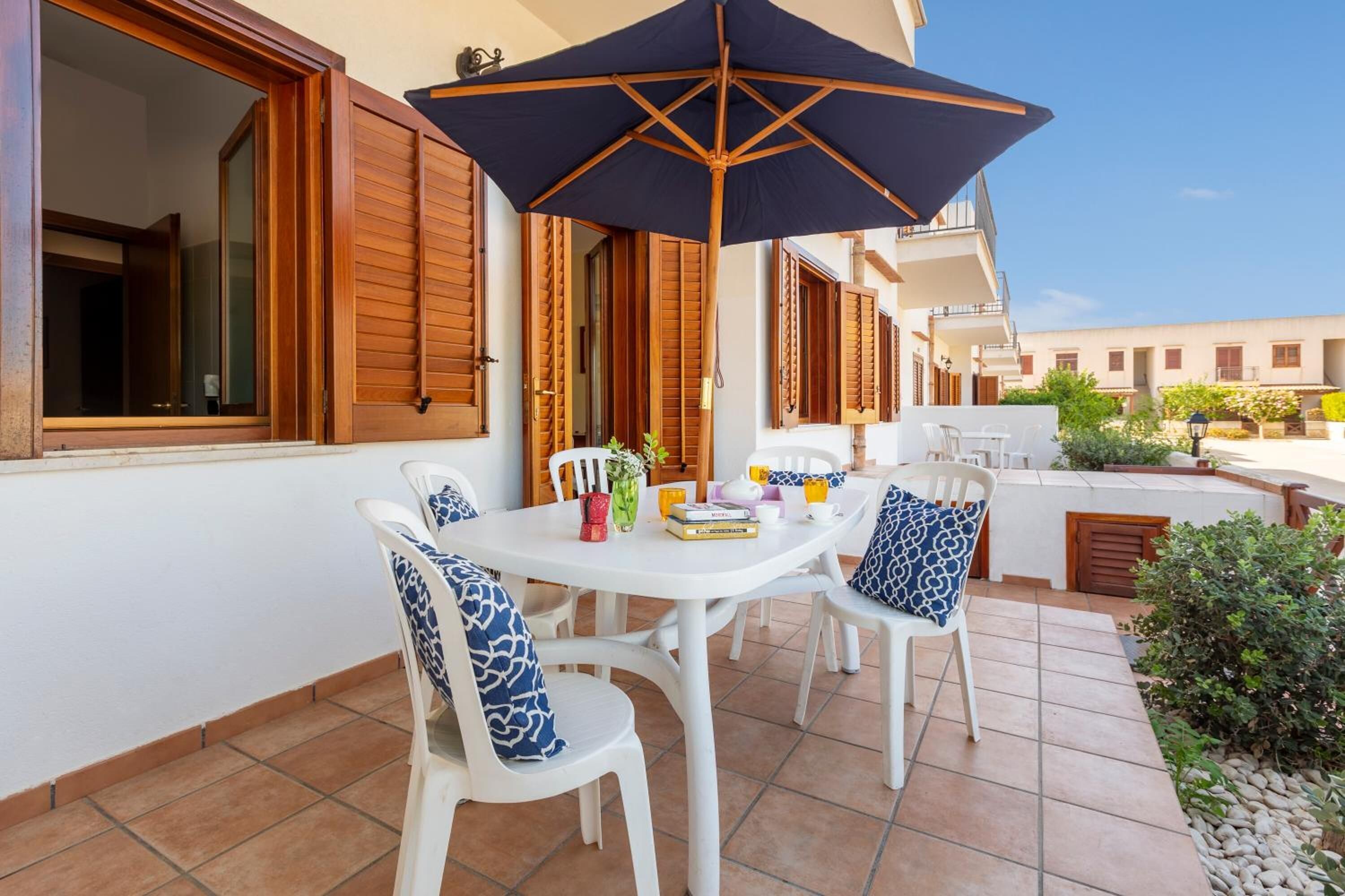 Un patio con una mesa y sillas blancas, un gran paraguas y contraventanas de madera en las ventanas.
