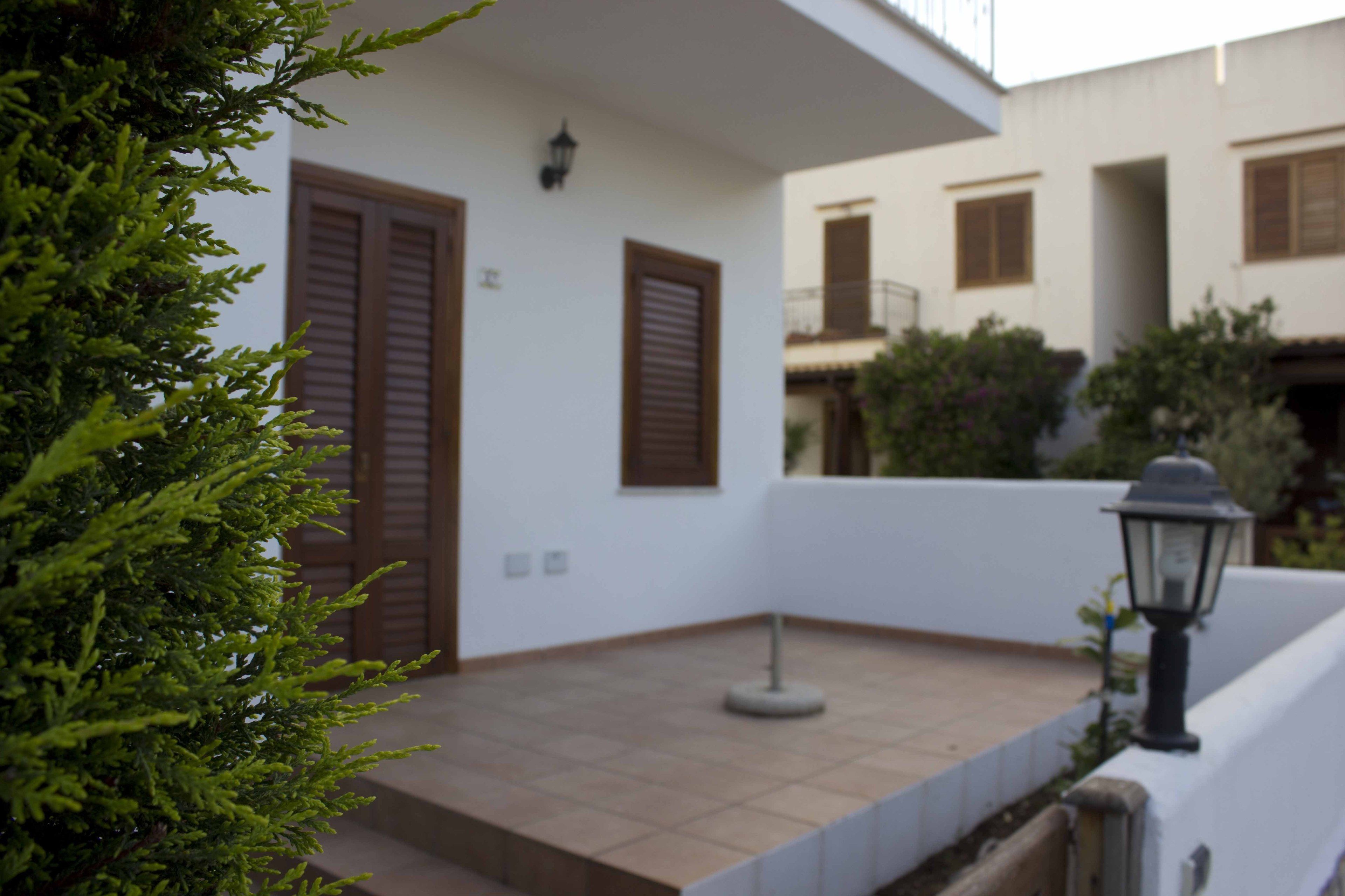 A white building with brown shutters, a tiled entrance, and a small garden area with a lamp post.