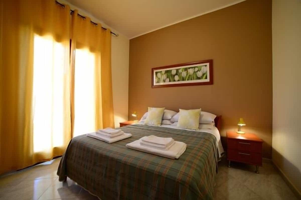 Une chambre d’hôtel avec un lit, deux tables de chevet, un tableau au mur et des rideaux à la fenêtre.