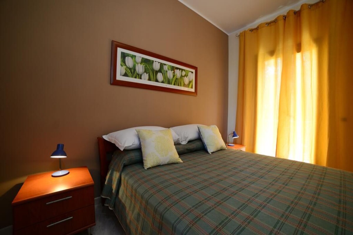 A bedroom with a bed, a nightstand, a lamp, a framed picture on the wall, and a window with yellow curtains.