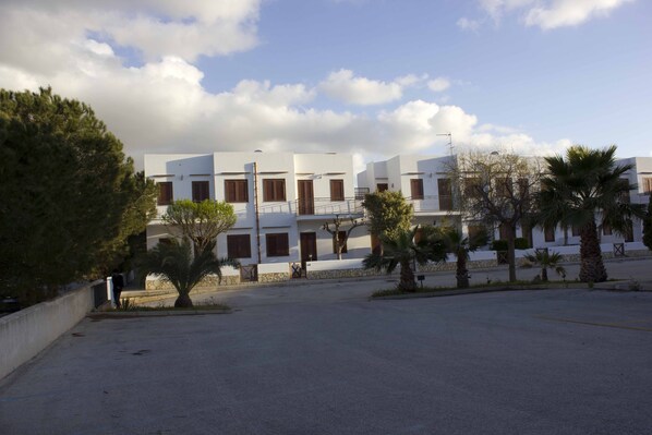 A row of white buildings with wooden shutters, a paved area in front, and palm trees.