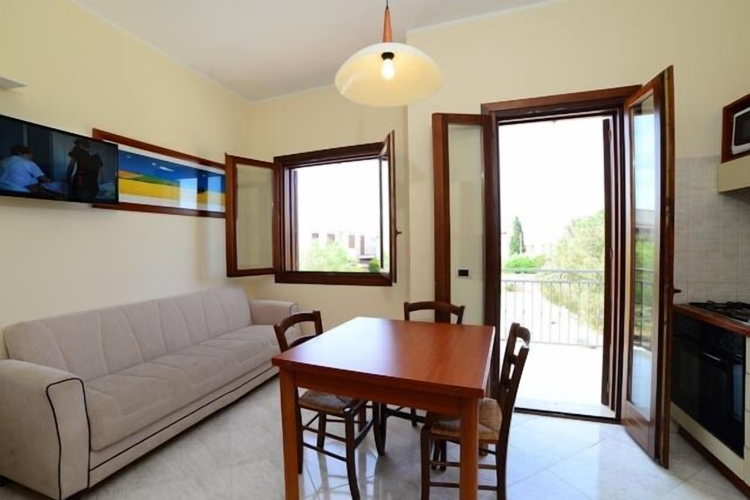 Una sala de estar con un sofá, una mesa de comedor de madera y sillas. Hay un cuadro en la pared, una ventana y una puerta que da a un balcón.