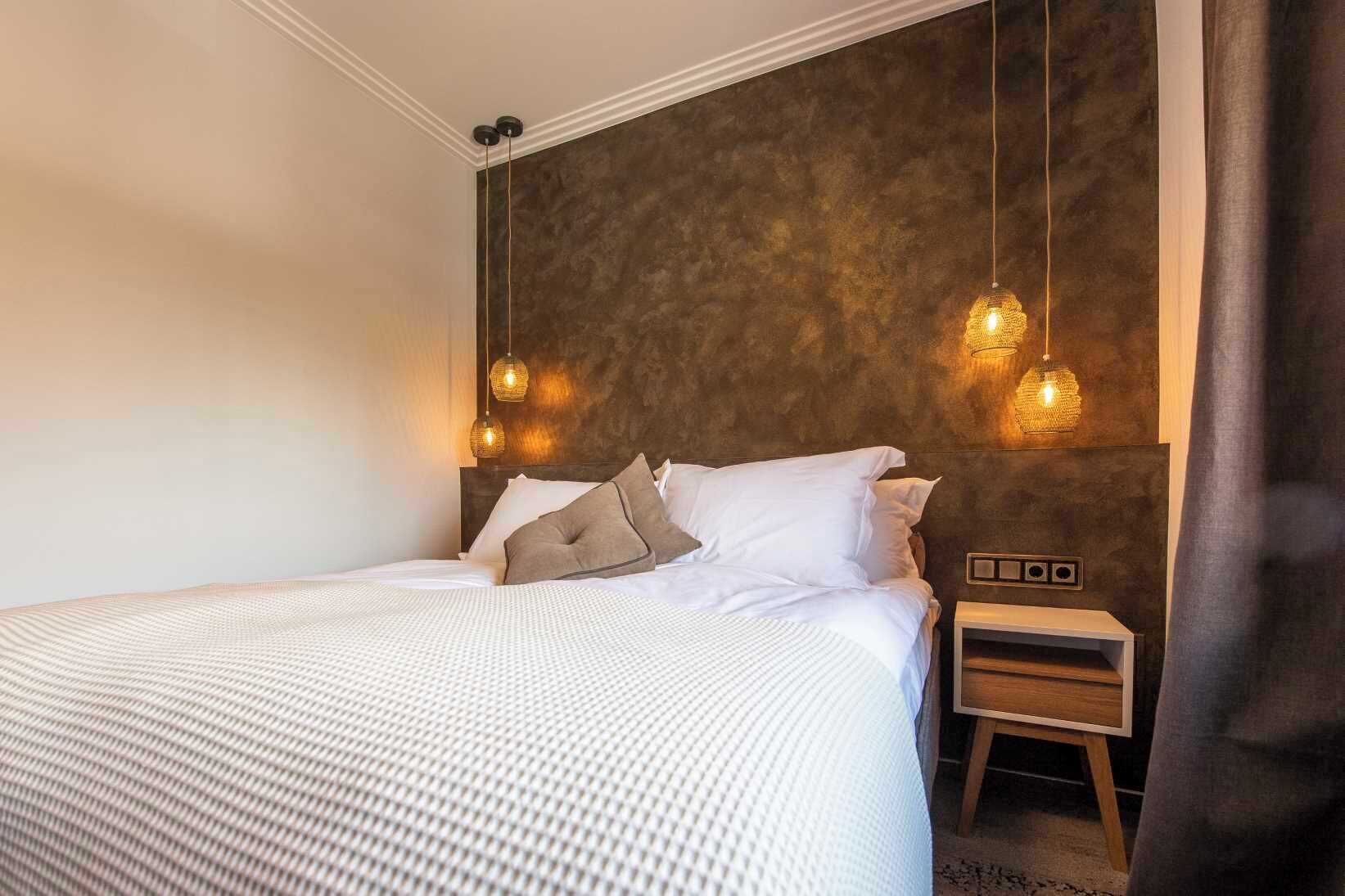A neatly made bed with white bedding and two pillows, a bedside table with a lamp, and a textured wall behind the bed.