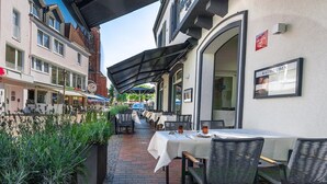 An outdoor dining area with tables and chairs, a covered walkway, and a building with a sign. - Ratshotel (Haltern am See)