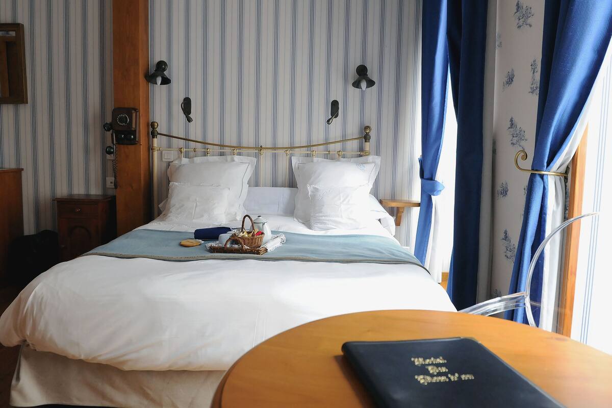 Una cama bien tendida con sábanas blancas, una mesita de noche con un libro y una ventana con cortinas azules.