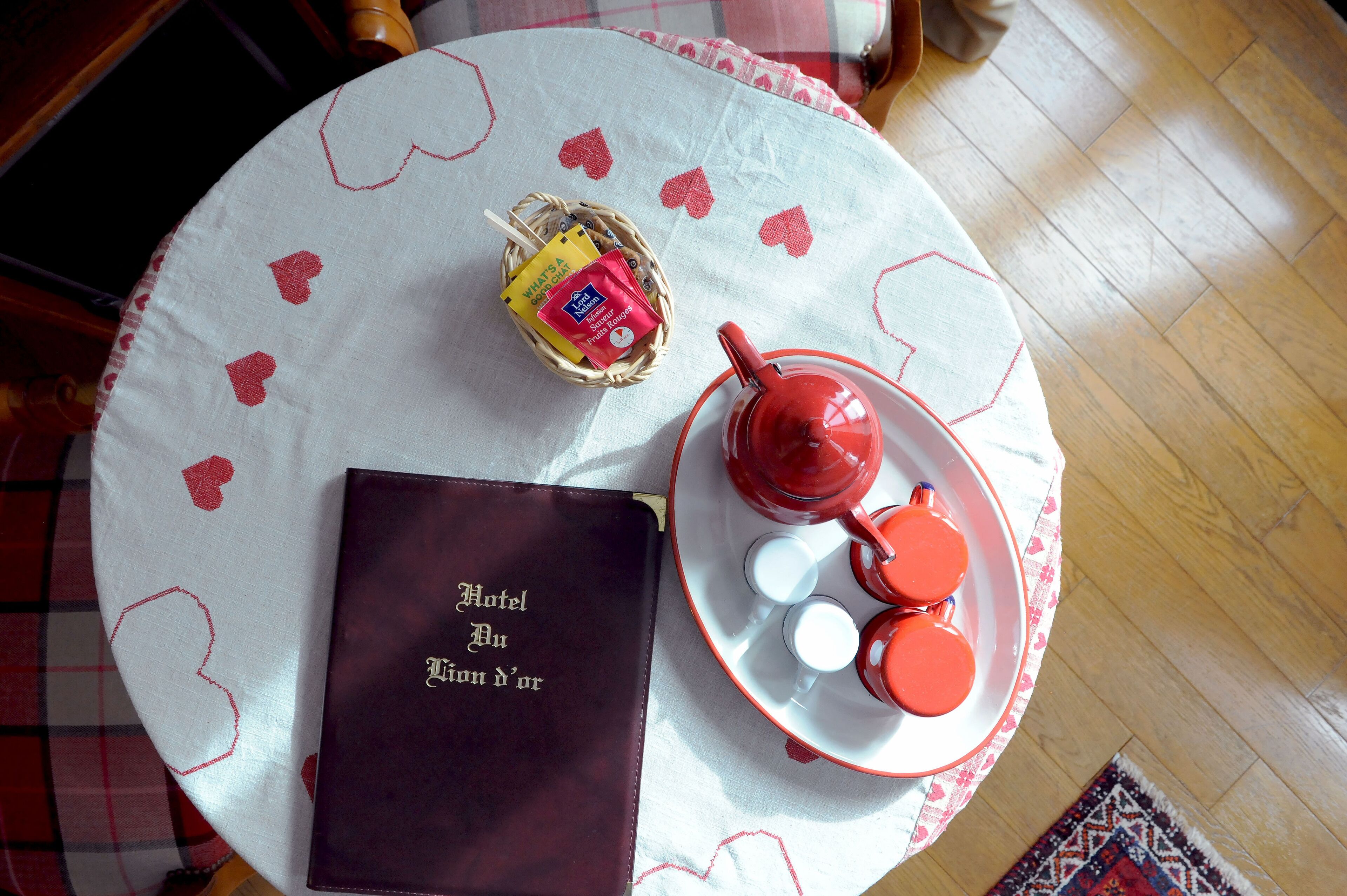 Une table ronde recouverte d’une nappe blanche ornée de motifs en forme de cœurs rouges, un panier en osier contenant un objet jaune et rouge, une théière rouge et un plateau avec deux tasses orange.