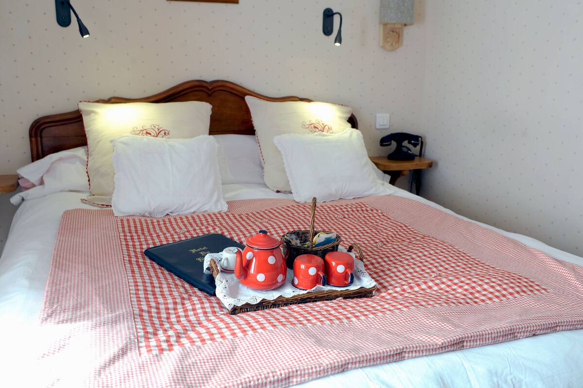 Una cama bien hecha con una colcha a cuadros, dos almohadas, una mesita de noche con una lámpara y una bandeja con tetera, tazas y un libro.