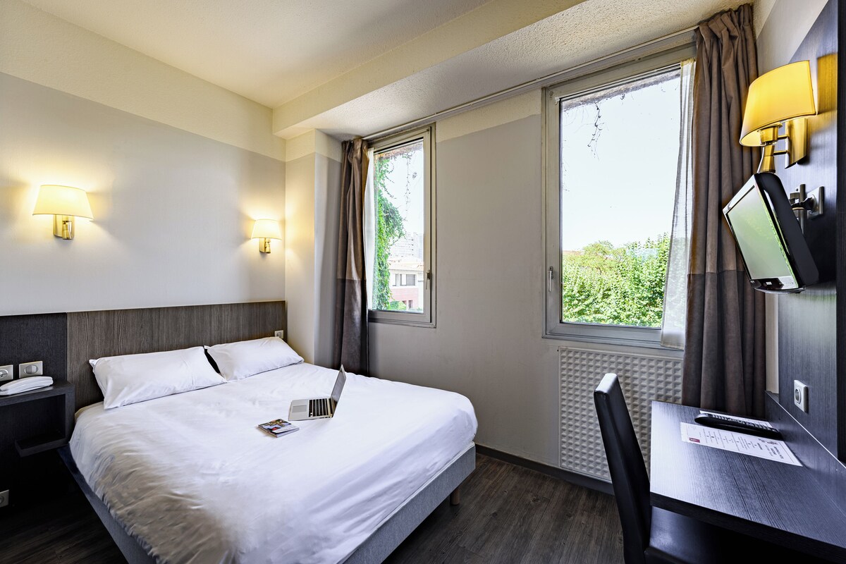 A hotel room with a bed, bedside table, lamp, TV, and a window with curtains.