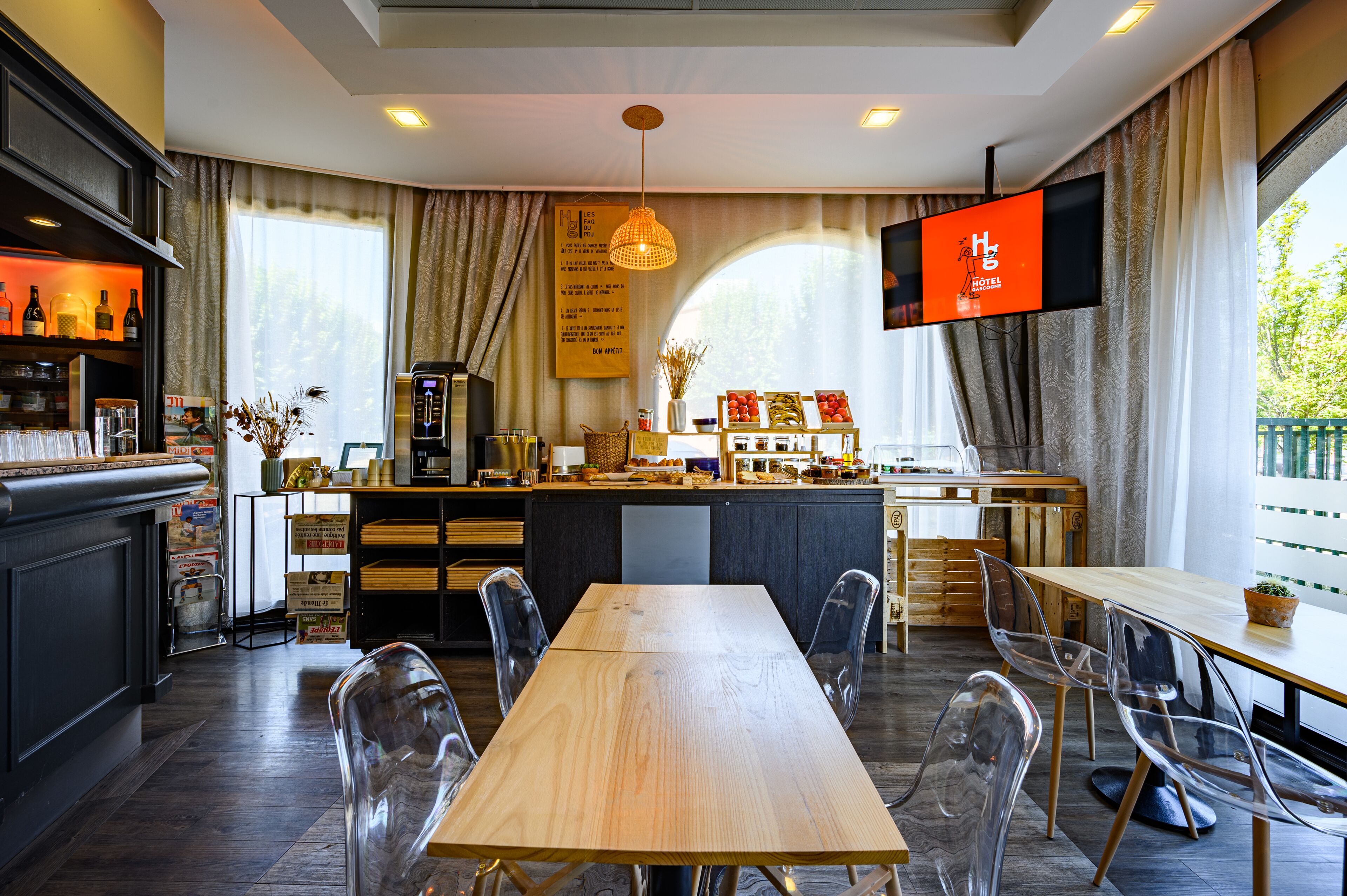 A modern hotel room with a wooden dining table, clear chairs, a bar area with bottles, and a flat-screen TV on the wall.