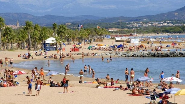 A crowded beach with people sunbathing and swimming, palm trees, and a lifeguard tower. - Hôtel Sable et Soleil (Fréjus)