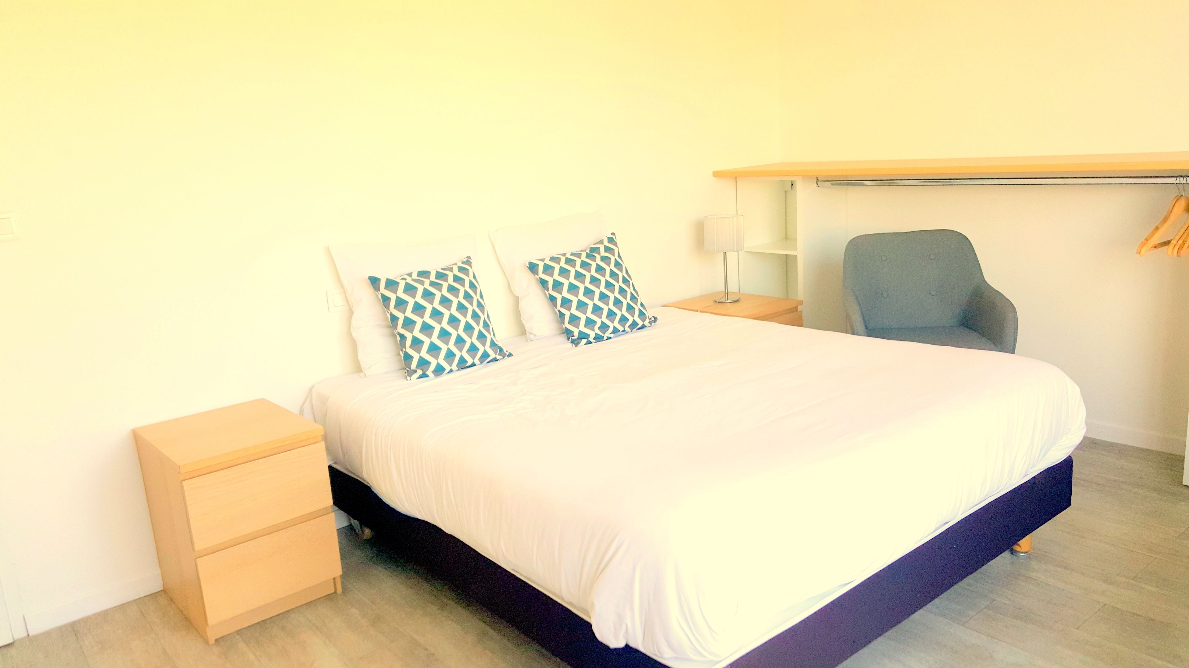 A neatly made bed with a white duvet and two patterned pillows, a small bedside table with a lamp, a chair, and a wooden nightstand.