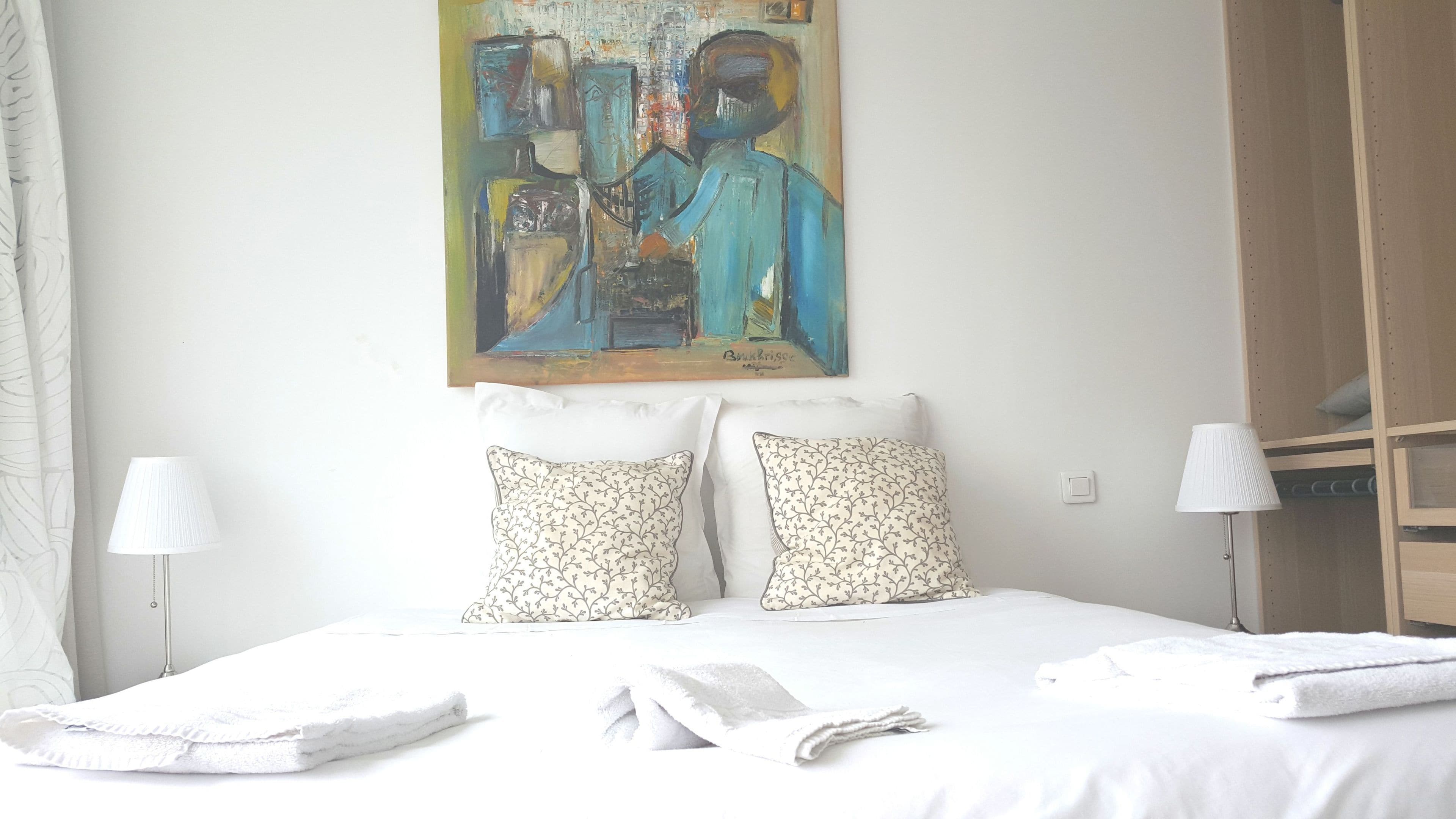Una habitación de hotel con una cama, dos almohadas, un cuadro en la pared, una mesita de noche y una lámpara.