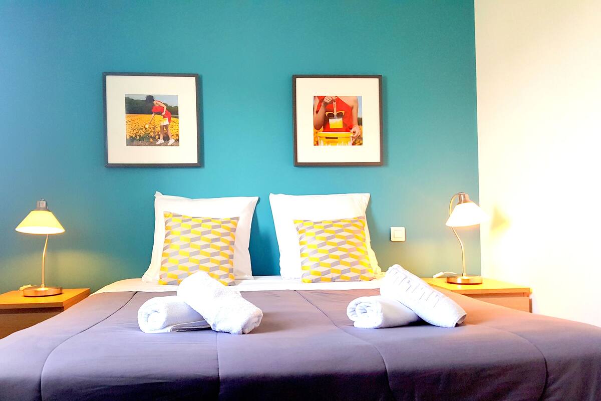 A hotel room with a bed, two pillows, two bedside lamps, two framed pictures on the wall, and two towels on the bed.
