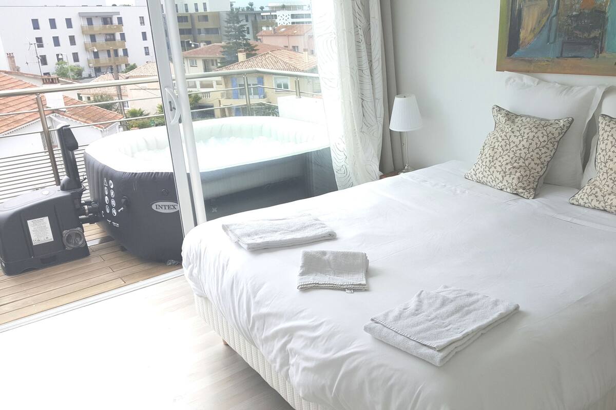 A hotel room with a large bed, two pillows, a window with curtains, a painting on the wall, and a view of buildings outside.