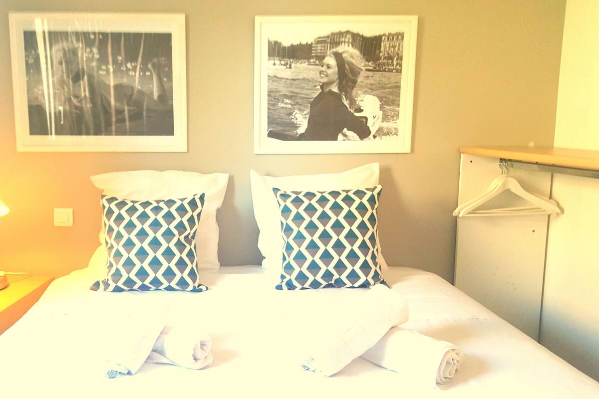 A hotel room with a bed, two patterned pillows, a nightstand with a lamp, and two framed pictures on the wall.