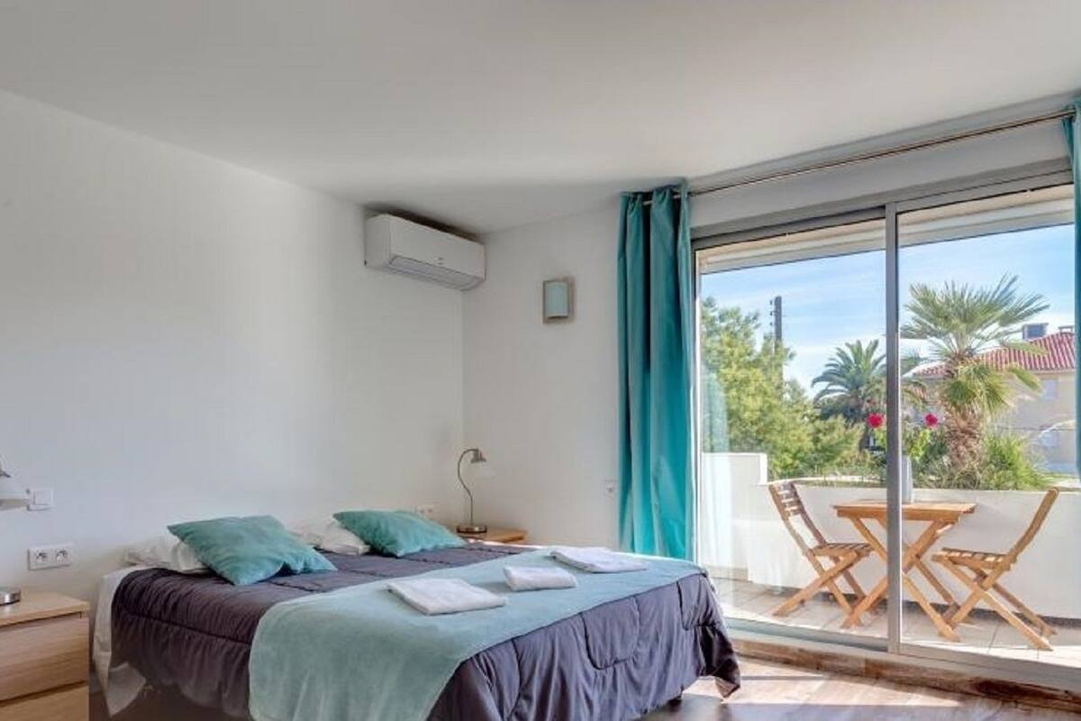 A bedroom with a bed, bedside tables, a desk, and a view of a balcony with outdoor furniture.