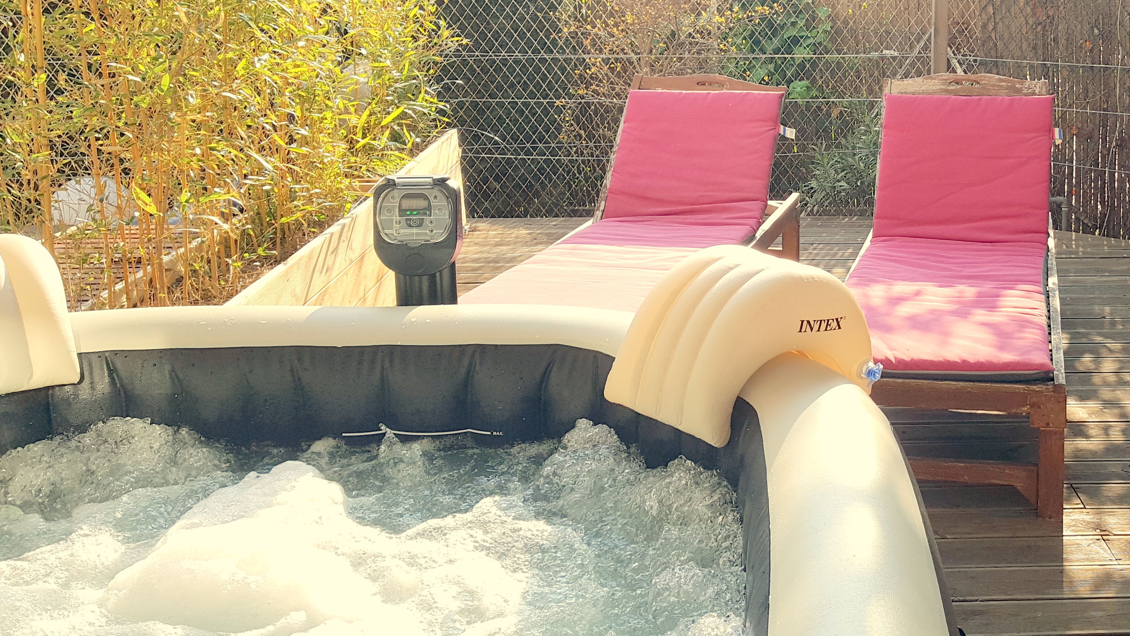 Un jacuzzi inflable con panel de control, rodeado por una terraza de madera y dos sillones reclinables rosados.